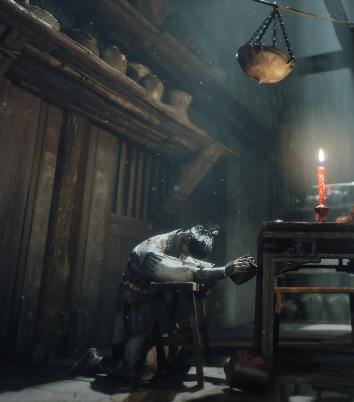 A figure kneels beside a small table with a lit red candle in a dim wooden room, a hanging bowl and stacked jars overhead.